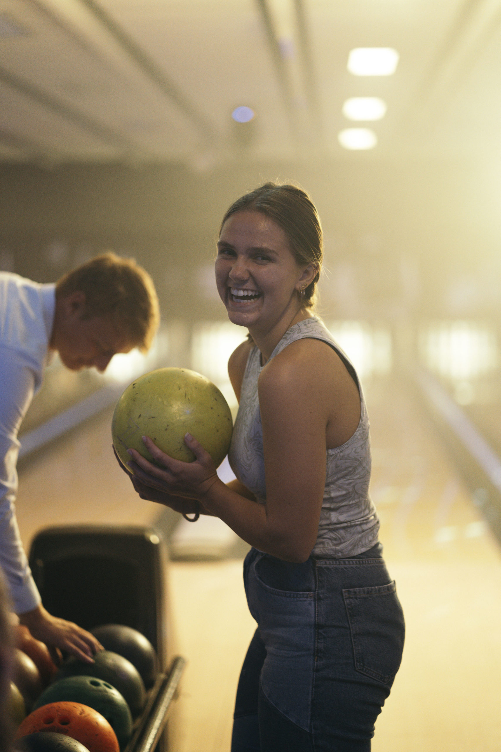 Pige kigger ind i kameraet, hvor hun griner og holder en gul bowlingkugle